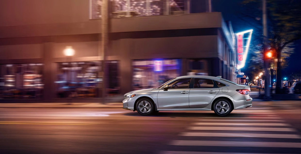 2025 Honda Civic Hybrid in silver driving through the city at night, highlighting its efficiency and modern style.